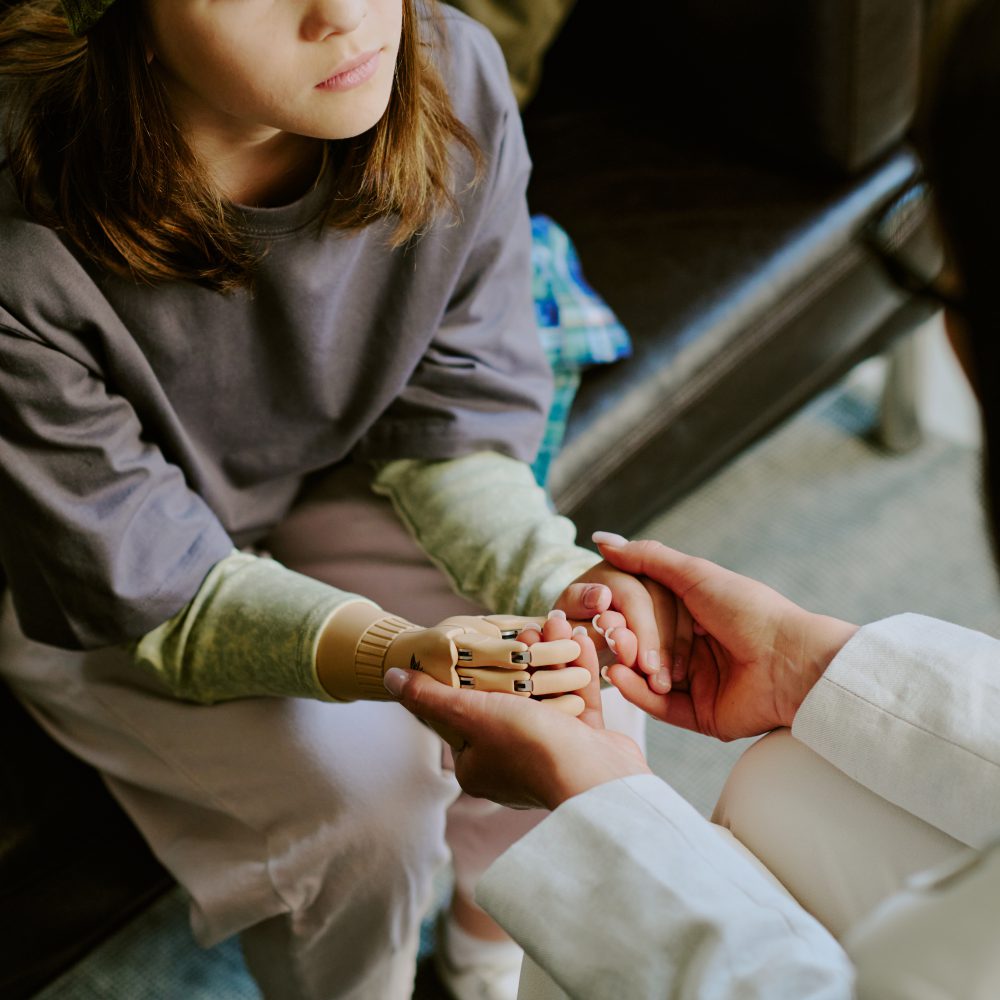 Child sitting on couch, receiving comfort from person holding robotic hand, reflecting support and technological advancement. Details emphasize childs thoughtful expression