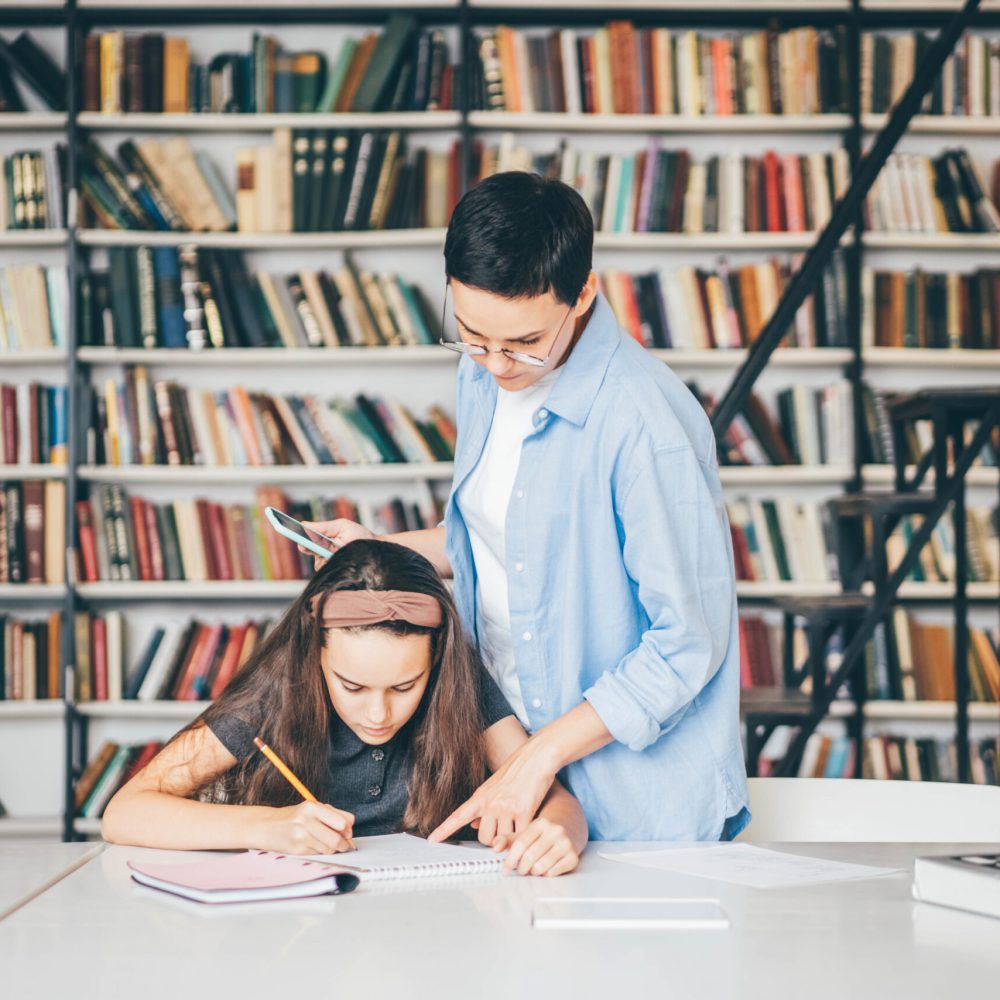 Teacher assisting girl with homework in library. Tutor working with middle school student.