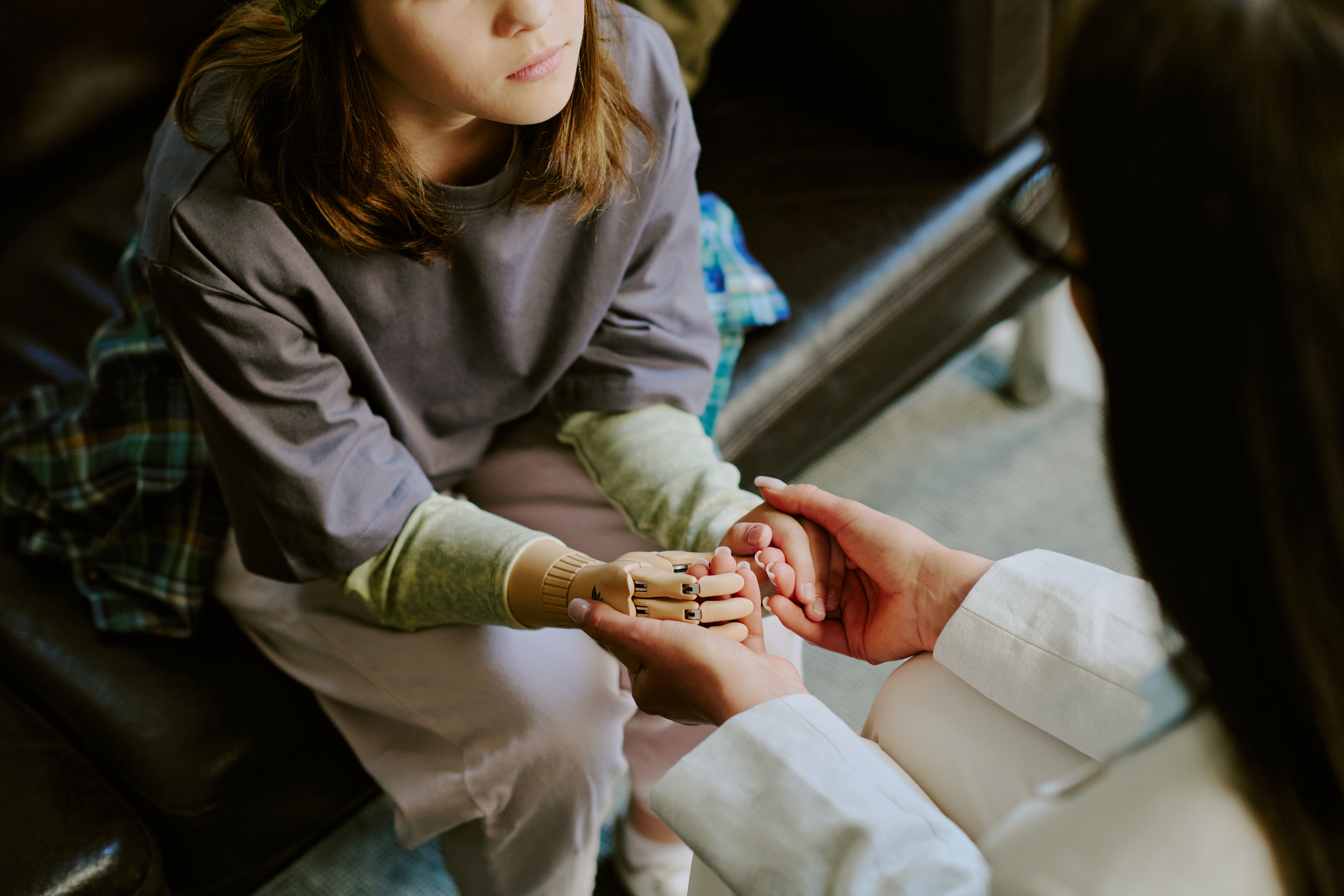 Child Receiving Comfort While Holding Robotic Hand Child sitting on couch, receiving comfort from person holding robotic hand, reflecting support and technological advancement. Details emphasize childs thoughtful expression